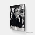 Black-and-white photographic canvas print of a woman in a dark jacket resting her chin on her hand, seated at an outdoor table with a checkered cloth and glass.