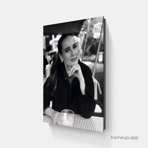 Black-and-white photographic canvas print of a woman in a dark jacket resting her chin on her hand, seated at an outdoor table with a checkered cloth and glass.