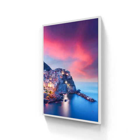 White-framed canvas print featuring a vibrant coastal village at twilight with pink and purple skies above blue waters.