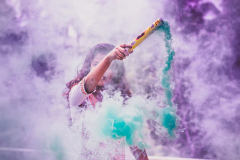 Person holding a colorful powder cannon amidst a cloud of vibrant purple and teal smoke.
