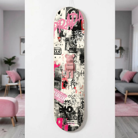 A pink bearbrick figure with ’prada’ branding sits centrally on a skateboard deck adorned with graffiti-style graphics, black and white street art elements, bold pink lettering including the word “prada,” and splashes of vibrant magenta.